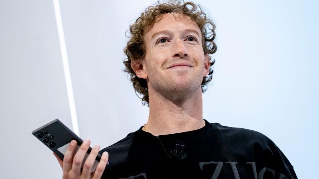 Mark Zuckerberg at a press event, smiling while holding a mobile phone.