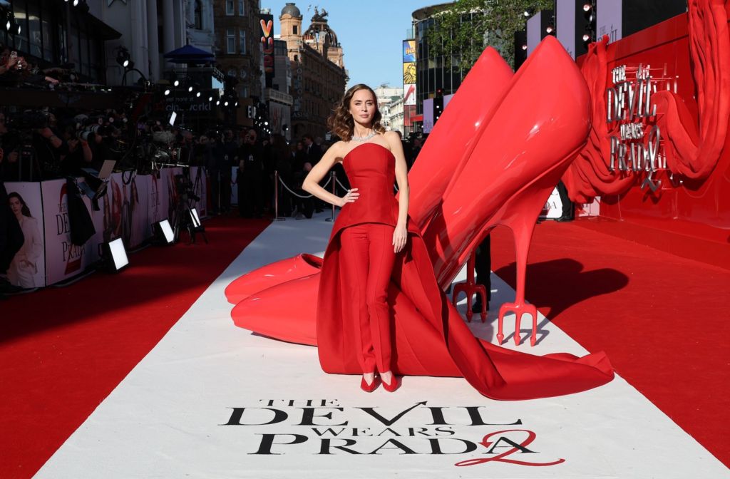 Emily Blunt poses at the red carpet premiere. She is standing next to a sculpture of a giant pair of red stilettos. 