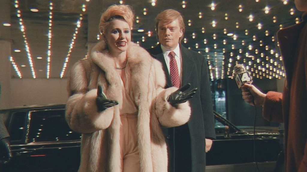 Maria Bakalova and Sebastian Stan as Ivana and Donald Trump in The Apprentice - Bakalova is wearing a fur coat and black leather gloves, Stan has a red tie, white shirt and black overcoat. They are being interviewed by a reporter.