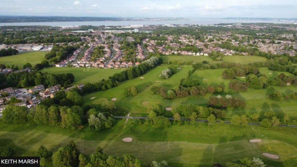 An aerial shot of the golf course, with numerous trees and holes seen and some housing to the left and behind the course, and the River Mersey in the distance