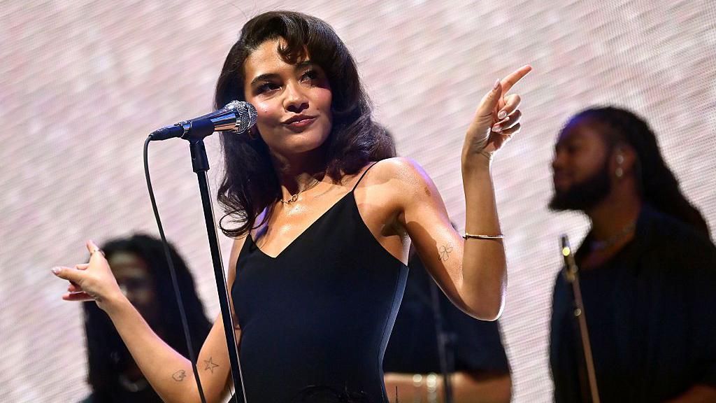 Olivia Dean wears a black dress and is performing on stage in front of a microphone. She is looking off to the side and pointing outwards with her hands.
