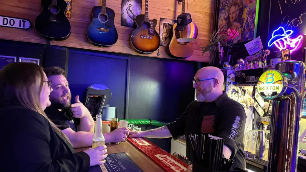Two people stand at Morgan's bar counter holding drinks while speaking with him. The bar surface has branded mats, straws, and drink taps visible. Behind the bar, shelves contain bottles, decorations, and a neon sign shaped like a hat. Several guitars hang on the wooden wall above the seating area.