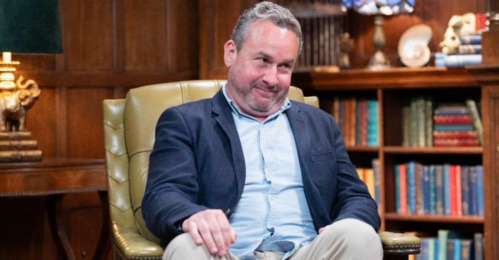 Hugo on The Traitors, sitting in an armchair speaking to Claudia