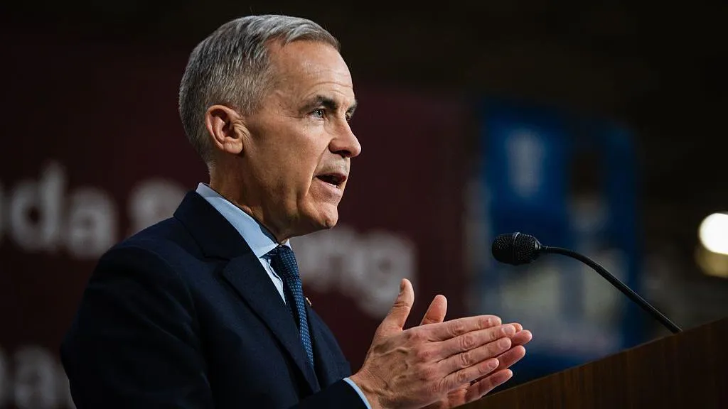 Head and shoulders image of Mark Carney viewed from the side. He is in front of a microphone and is holding his hands close together as if about to clap. 