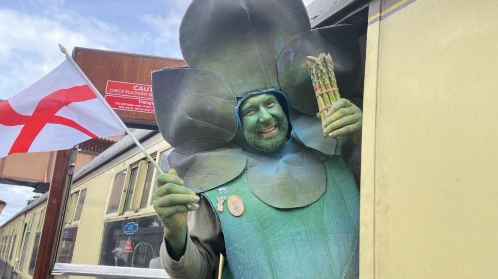 A man in an asparagus costume holding a bundle of asparagus in one hand and the flag of England in the other whilst hanging out of a train