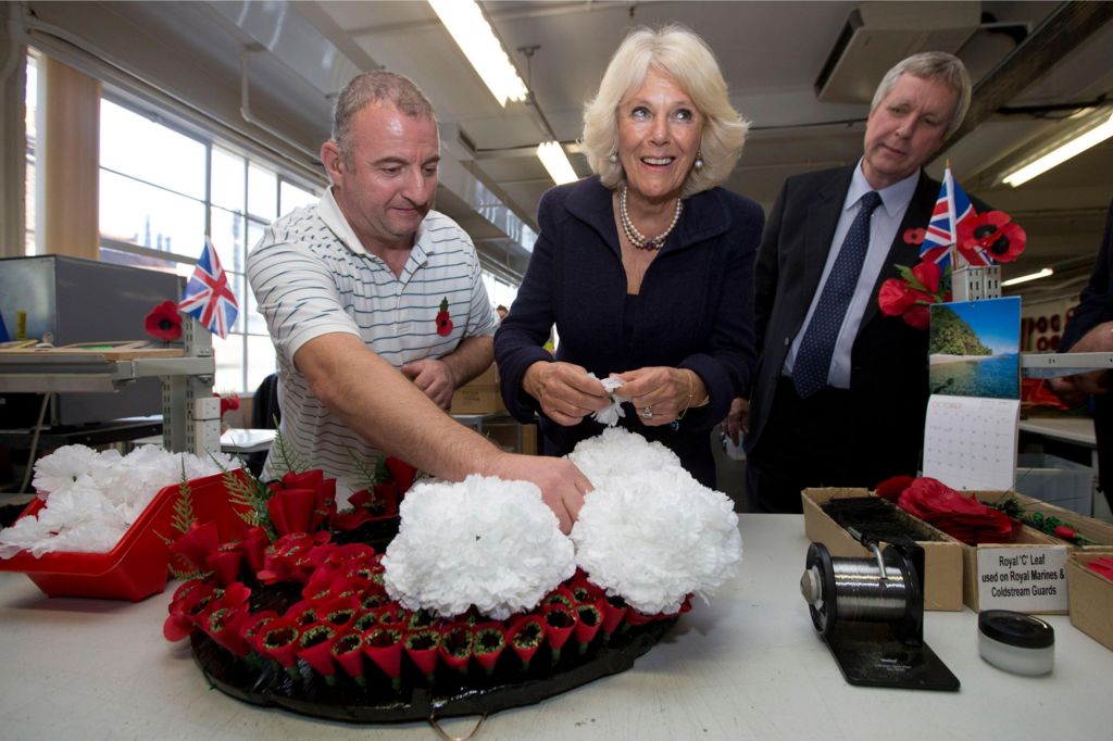 Man in striped shirt holds white flowers on top of red poppy wreath as woman with pearls smiles and prepares to add flowers