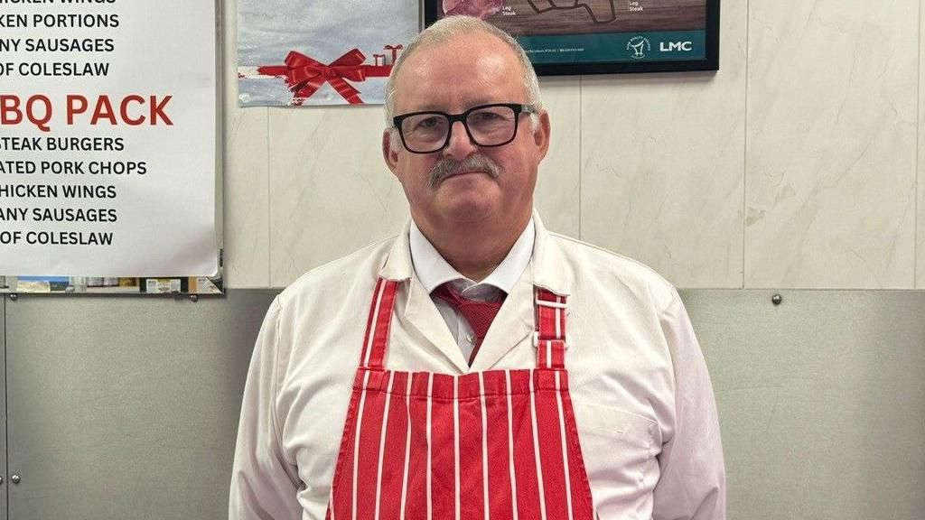 Portadown Butcher Phillip Freeburn stands in his red stripey apron in front of his shop. He has white hair, a small moustache and glasses.