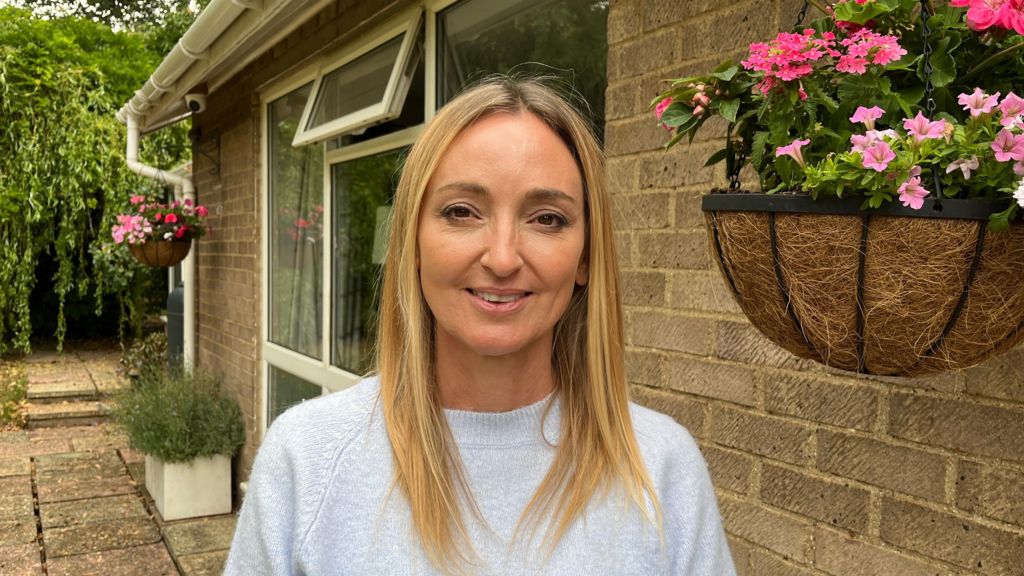 Claudia Pixley, a woman wearing a light blue top with long blonde hair, stands outside her home with a hanging basket next to her.