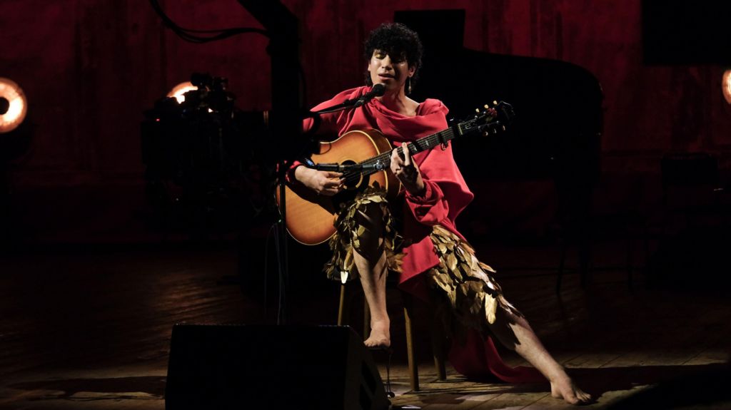 Jacob is sitting on a stool playing a guitar and singing into a microphone. They are performing barefoot in a pair of golden-feathered trousers and a red shawl.