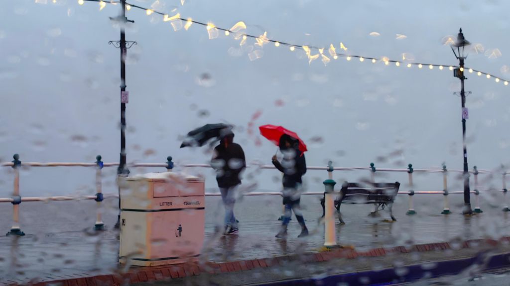 A photo taken through a window pane blurred with rain, showing two people walking under umbrellas