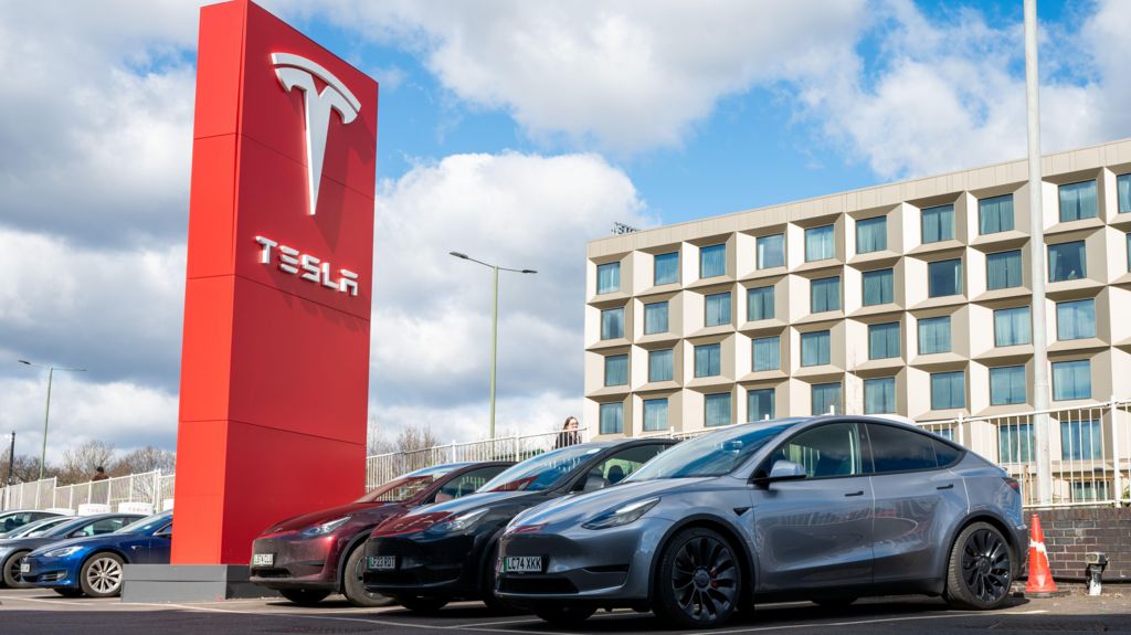 Tesla cars sit parked outside at a dealership in London. They are parked beside a red Tesla sign.