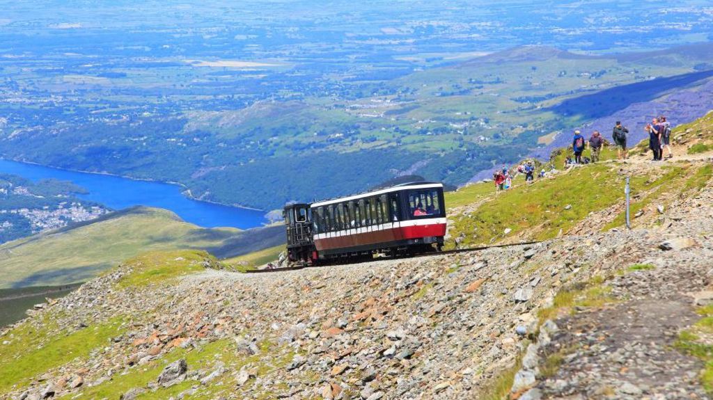 Train on the Snowdon Mountain Railway tracks on Yr Wyddfa - Wales' highest peak, with walkers off to one side and views looking out across Llanberis and Llyn Padarn lake