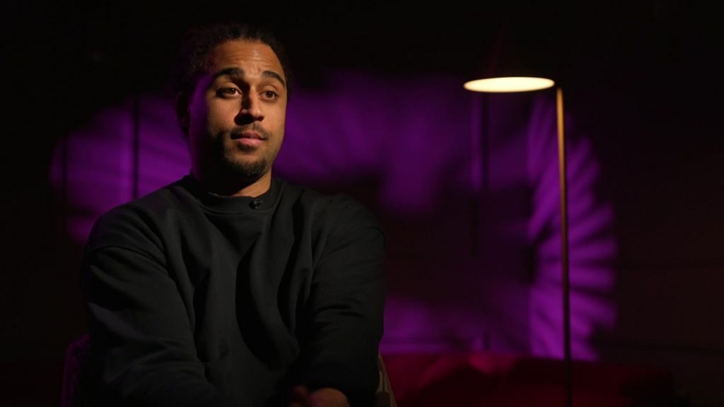 Sebastian Wray sits in a studio interview, wearing a dark sweatshirt, with purple lighting and a floor lamp behind him.