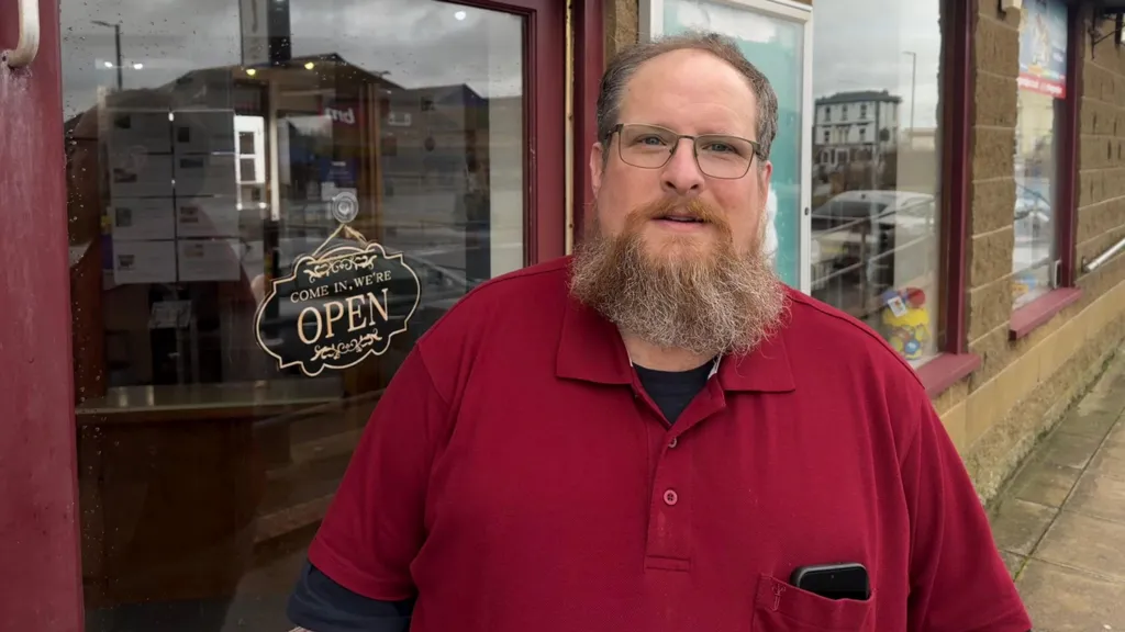 Simon Gready is standing outside a shop door wearing a red shirt. He has a beard and is wearing glasses.