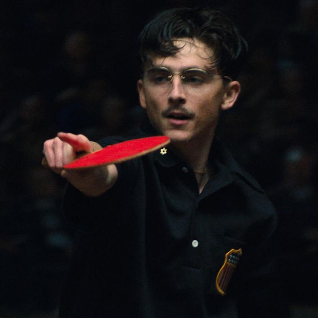 A still from Marty Supreme showing Timothee Chalamet's character playing table tennis, pointing with a table tennis bat in one hand