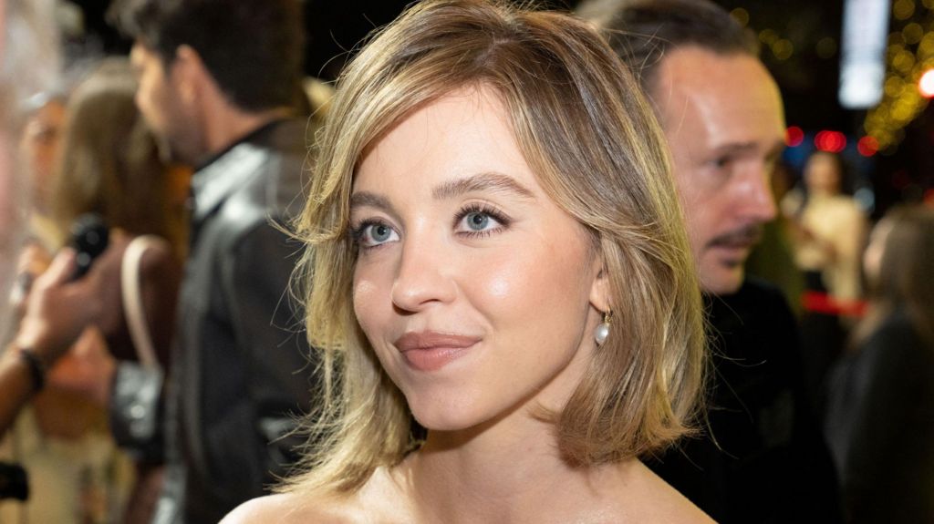 Sydney Sweeney in a strapless dress on a red carpet. She is wearing pearl drop earrings and has her dark blonde hair in a bob.