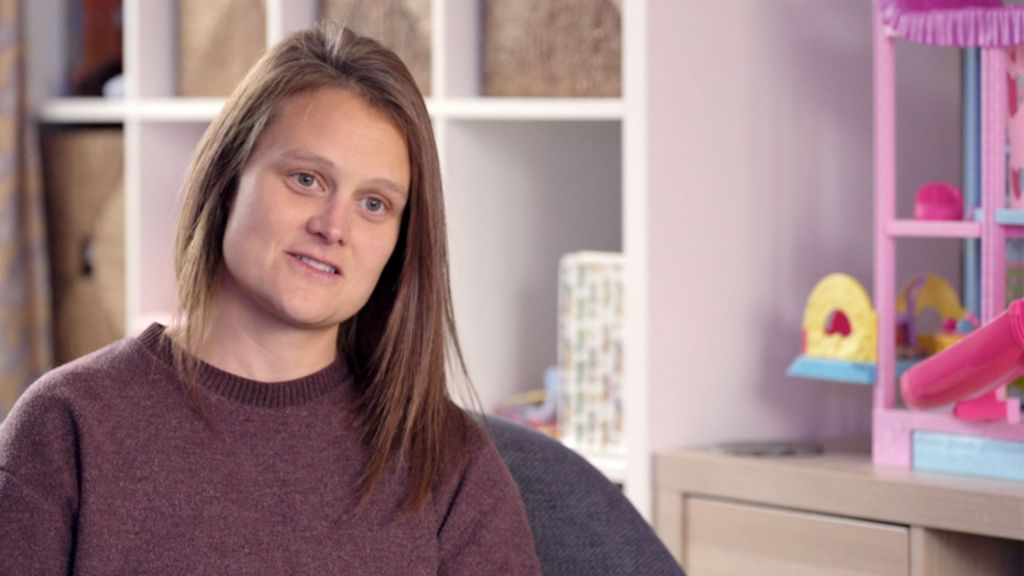 Becky has long brown hair and wears a dark purple top. Behind her is a Barbie doll house and a large white shelving unit.