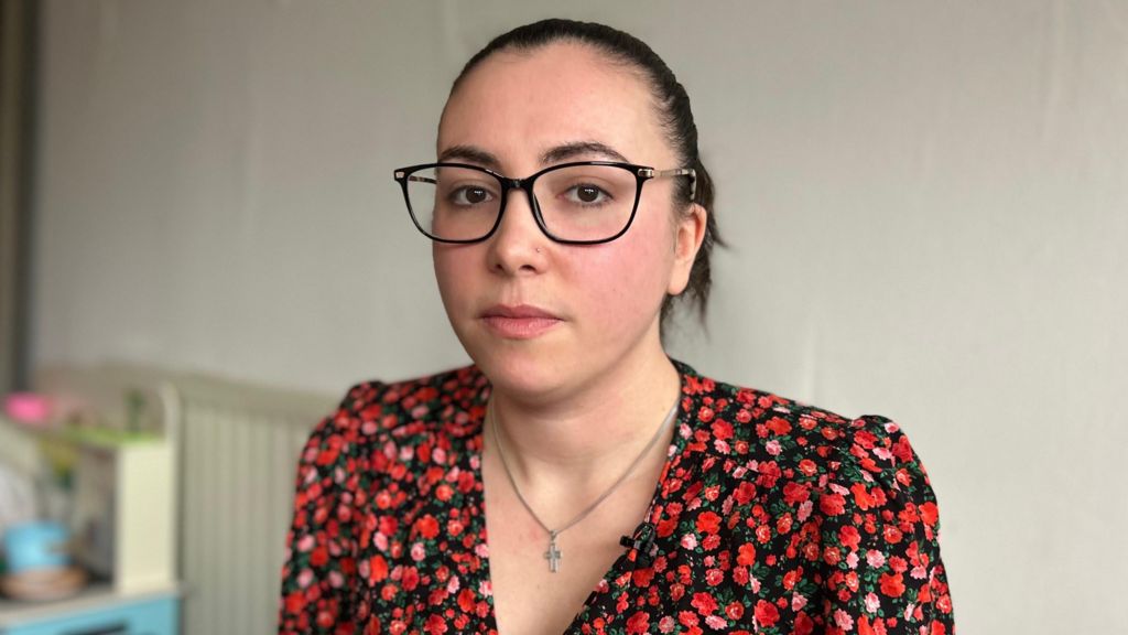 A woman sitting in her living room stares into the camera lens. She has brown hair scraped back in a ponytail, and is wearing a floral red top, a cross necklace, and black-rimmed glasses. 