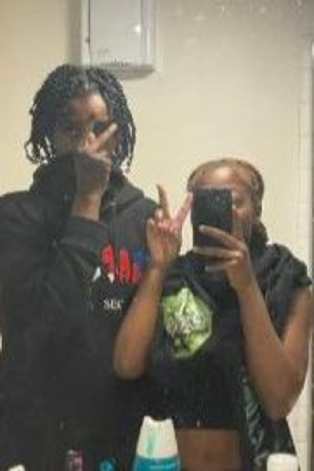 Teenage boy dressed in black covers his hands with his face alongside teenage girl holding up peace sign with her mobile covering her face.