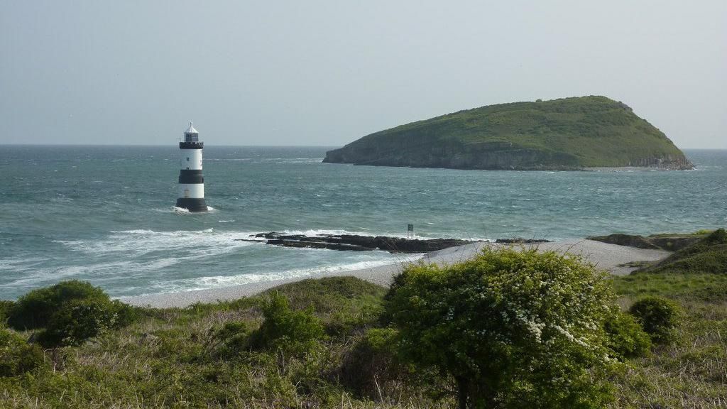 A lighthouse appears on a small island between the beach at Penmon and a larger island, Puffin Island, in the distance.