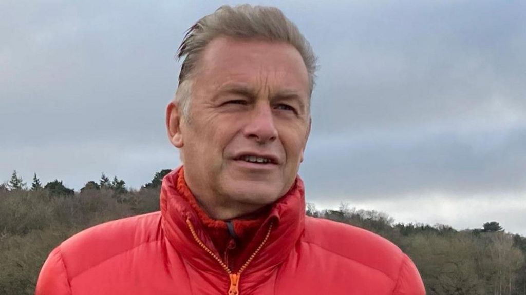 Chris Packham looks off camera during an interview, he is wearing a red puffer jacket zipped up with a red fleece jumper underneath, there is woodland in the background