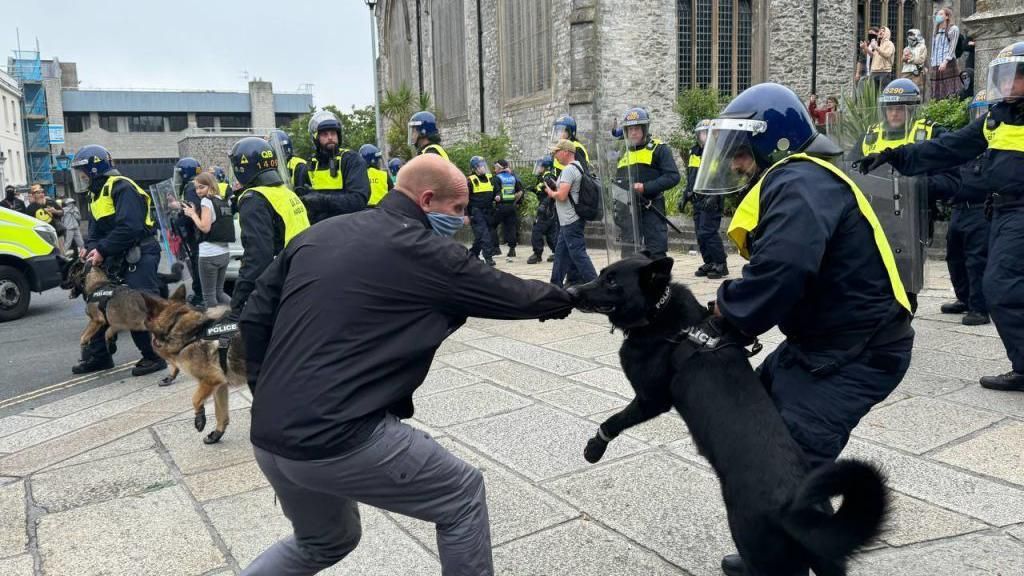 A protester and police dog appearing to lunge at each other with police officers in riot gear nearby