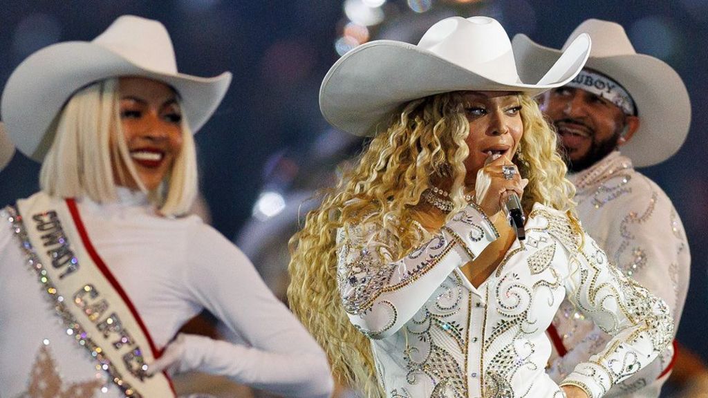 Beyonce in a white cowboy hat and jewelled white suit singing into a microphone flanked by dancers in similar outfits, performing at half-time furing an NFL game on 25 December 2024