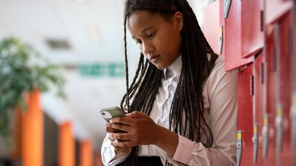 A teenager looks down at a mobile phone. She is wearing a white shirt as part of a school uniform with long dark brown hair. She is leaning against some red lockers. 