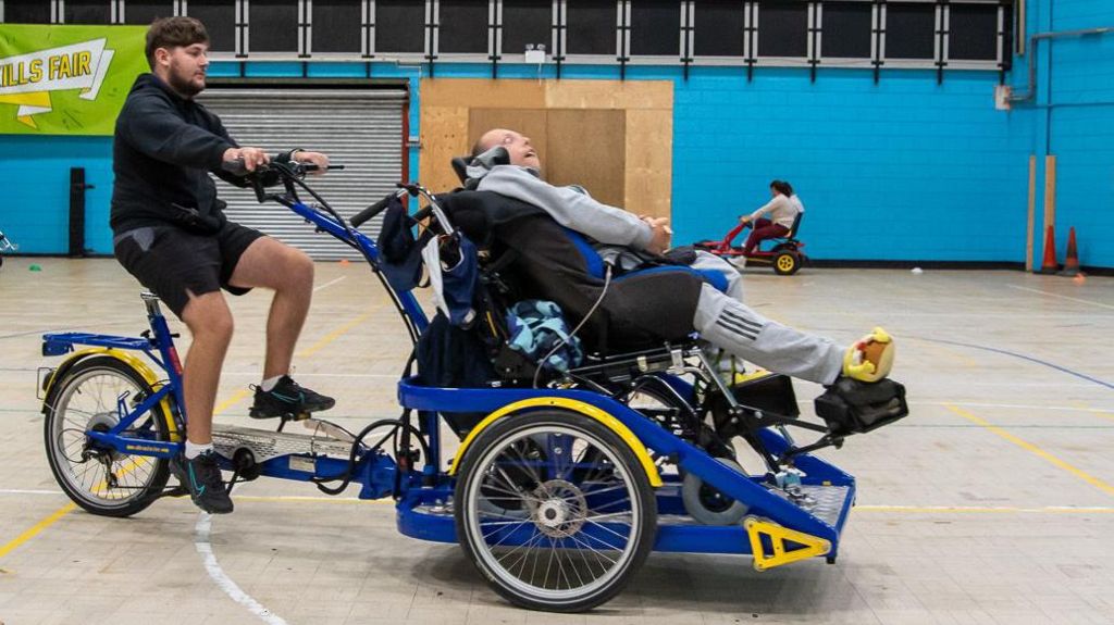 Two men ride on a large, blue adapted bicycle. It has one wheel and a standard bike seat at the rear, where a man in a black top and shorts sits and pedals. At the front, two wheels and a large steel structure support a recumbent seat, where a man in a grey top and jogging bottoms sits.