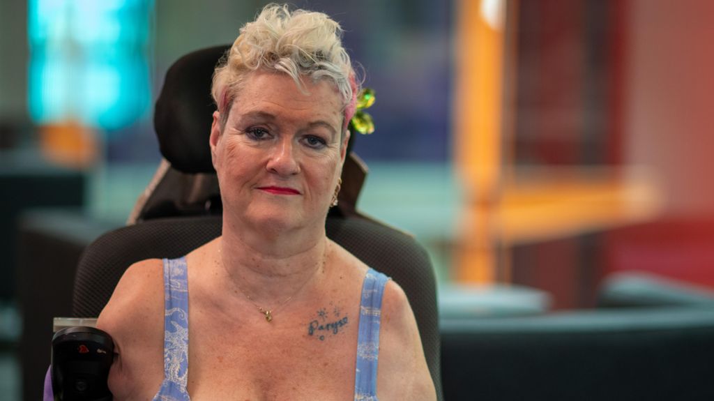 Alison Lapper is staring directly at the camera. She is wearing a blue outfit and has a pink and green hair clip in her blonde hair. Her son's name is tattooed above her heart. 