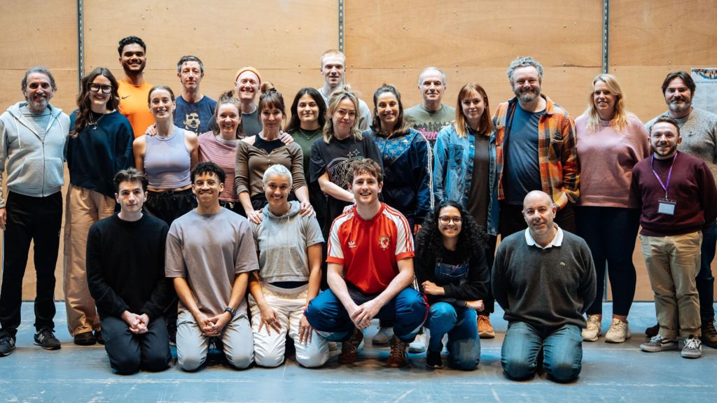 A group of more than 20 people pose together in rows in the rehearsal room