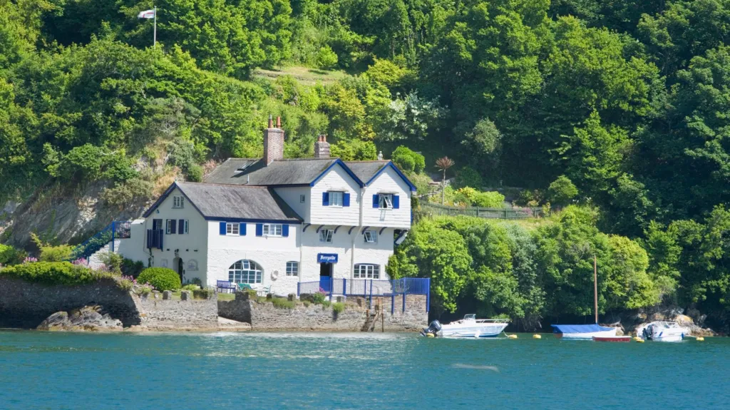 The old boatyard at Bodinnick is a large white wharf-style building overlooking the river Fowey. It is set in woodland and there are small boats moored in front of the building in the clear blue water. 