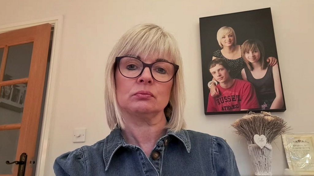 Sharron Huddleston, with blonde hair and glasses, looks at the camera with a forlorn expression. There is a photo of her with her two children on the wall behind her.