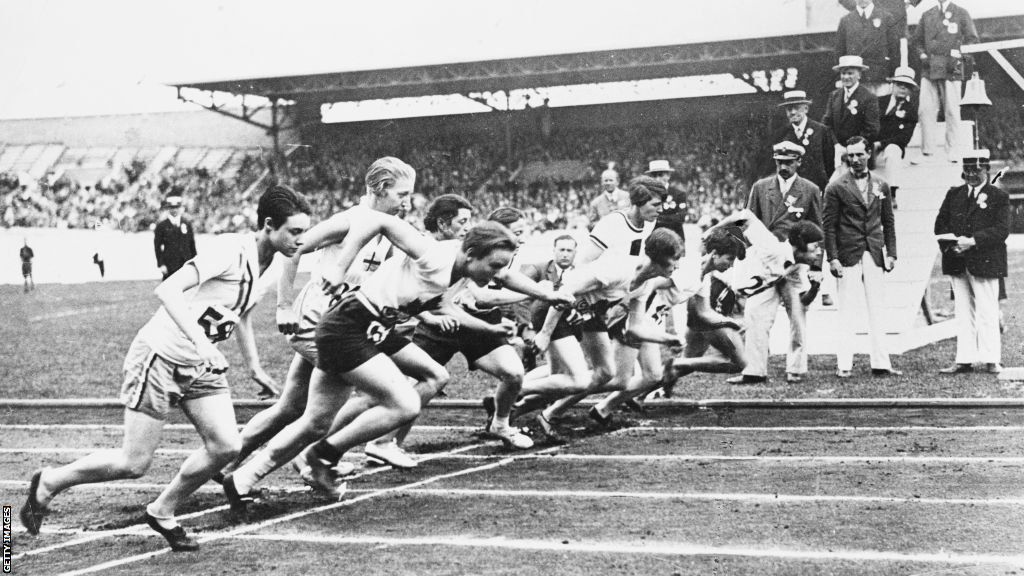 A group of runners sprint off the line, watched by suited male officials