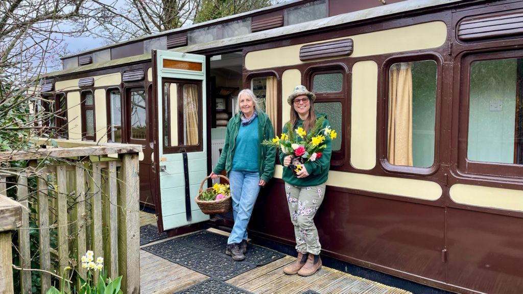Lizzy and Lil, two older women, standing outside an old train carriage which has been converted into self catering accommodation. Lizzy is holding a bunch of daffodils and Lil has a wicker basket of more flowers in her hand. 