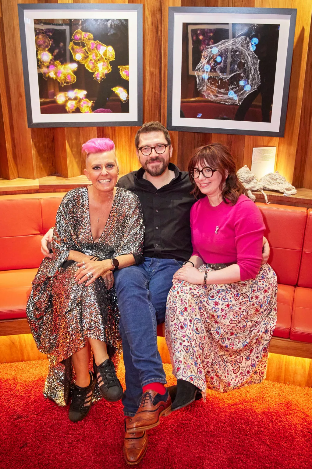 Three people on soft seating. A man in blue jeans and black shirt sits between a woman (right) with a floral skirt and pink top. On the left is a woman in a shiny, glittery dress. She has pink hair