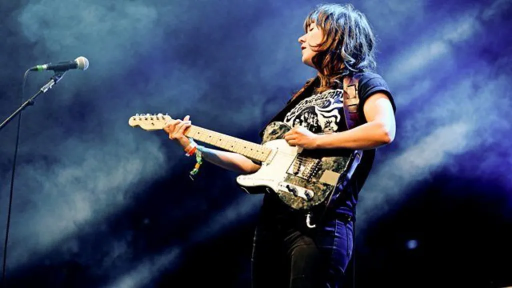 Dark haired woman on dimly lit stage set playing black and white electric guitar. She wears a black graphic t-shirt and jeans and has a shaggy hair cut.
