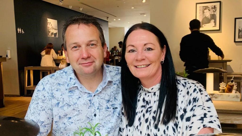 Dave and Lorna Ellard smiling together at a restaurant table. Dave has short black hair and is wearing a blue and white Hugo Boss shirt. Lorna has long jet-black hair and is wearing a white top with navy spots.