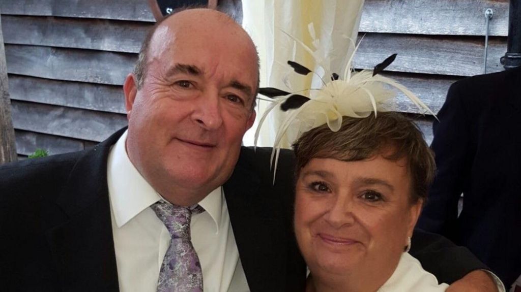 Rodrick Lodge wearing a suit has his arm around his wife Pauline who is wearing a black and white fascinator. They are both smiling and looking directly at the camera. 