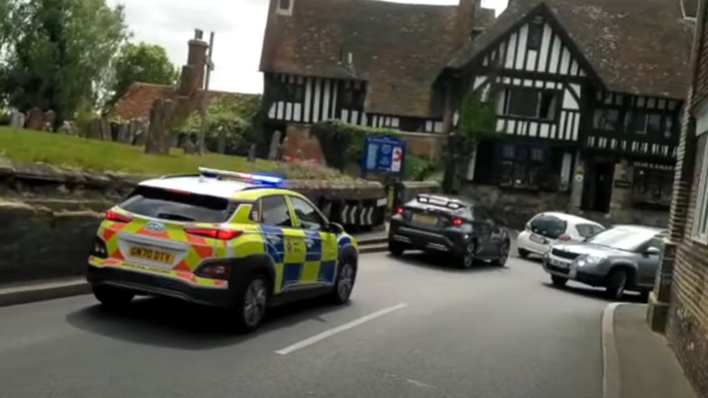 A police car with flashing sirens sits behind traffic stuck at a narrow corner