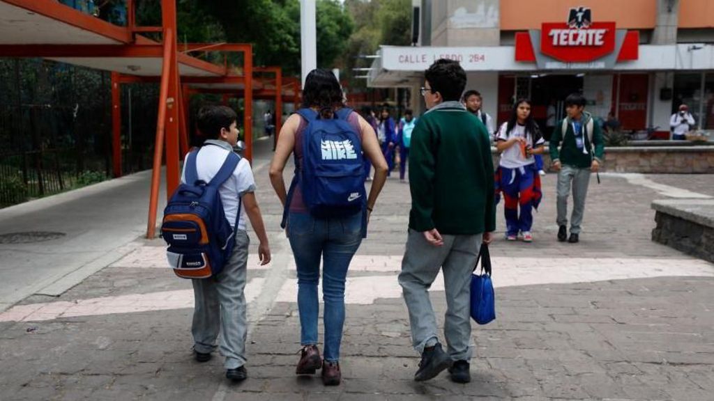 A mother picks up her children from an elementary school in Mexico City. She is flanked by a smaller boy, who is carrying a backpack, and a bigger boy, who is carrying a bag in his hand. Other children are walking towards them.