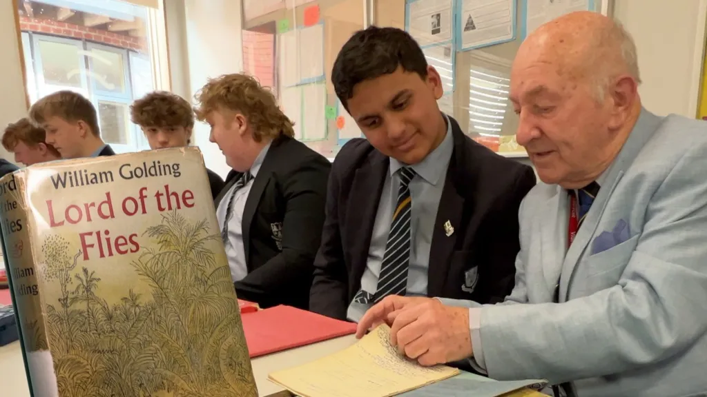 Mick (right) sits with current pupils of the school. Mick is wearing a light blue suit and a tie, and is looking through an old text book with a pupil who is smiling. Other pupils, all wearing black blazers, blue shirts and blue striped ties, are in the background. In the foreground there is a copy of Lord of the Flies, with a jungle scene on the cover. 
