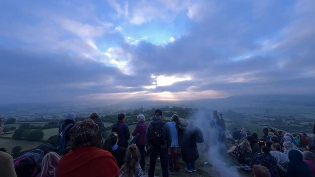 A view of the sun rising over the horizon at Glastonbury Tor. There are clouds in the sky but the sun is bursting through