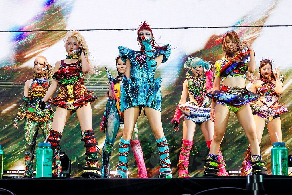 Members of XG are on stage in brightly-coloured costumes, holding microphones as they prepare to play at California's Coachella Festival