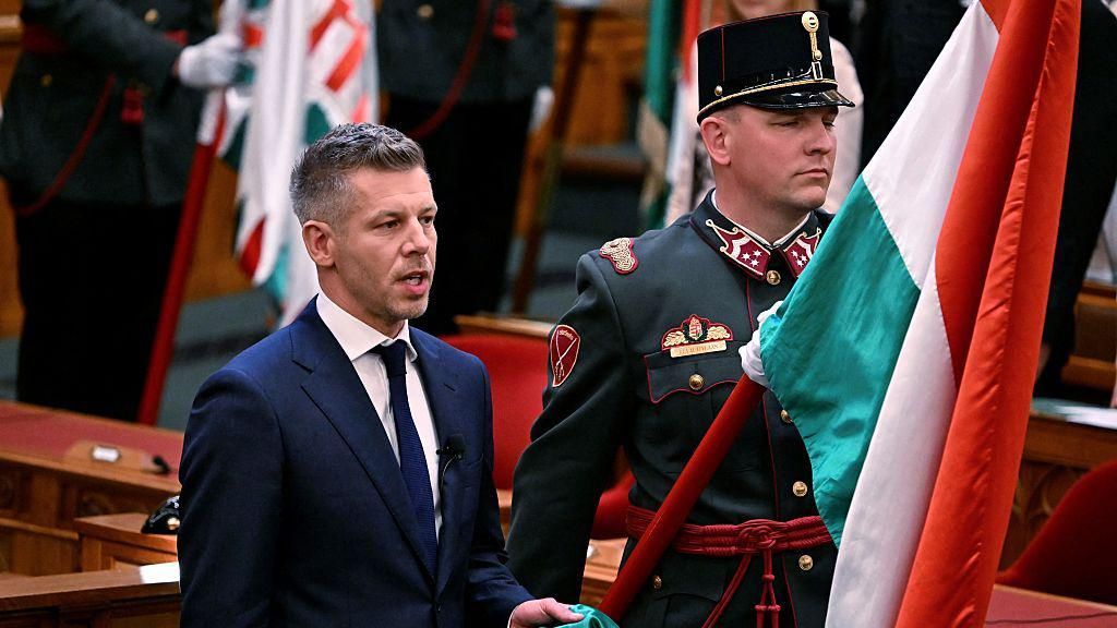 Peter Magyar is sworn in at the Hungarian parliament in Budapest on 9 May 2026. He is standing on the left and looking right, while beside him a man in uniform holds a Hungarian flag.