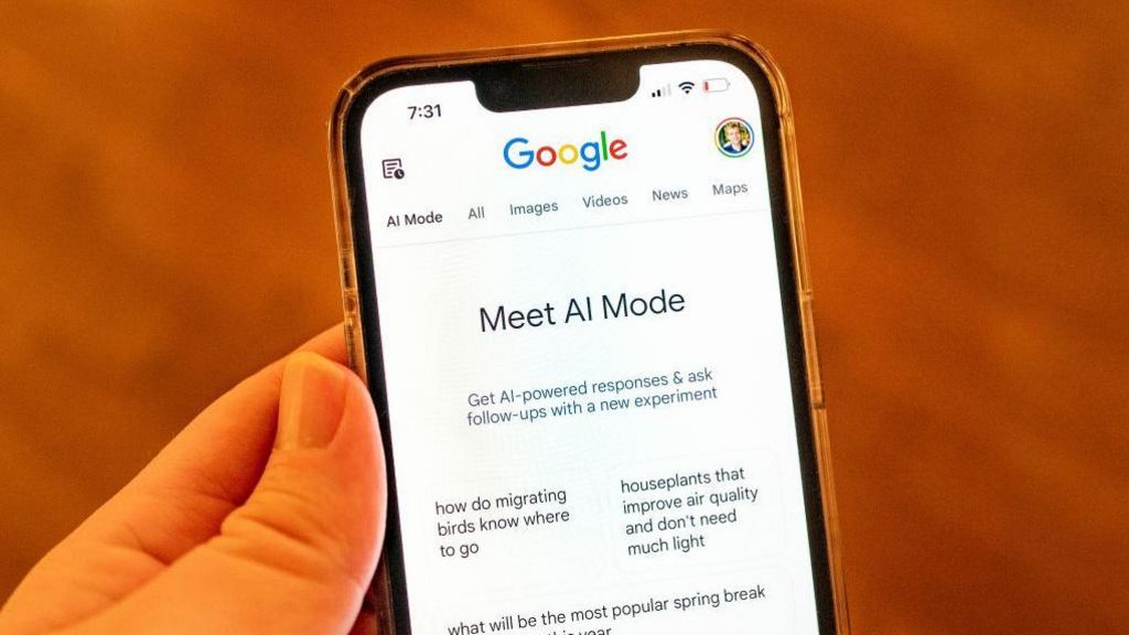A hand holding a mobile phone. On the screen is displayed Google across the top and then in the centre "Meet AI Mode". Below that there is text explaining that this tool will provide AI-powered responses and allow the user to ask follow up questions. Below that are example queries: How do migrating birds know where to go, and Houseplants that improve air quality and don't need much light.