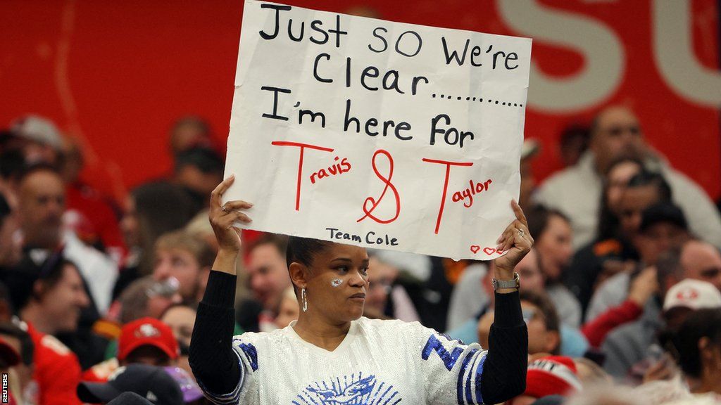 A fan holds a sign that says 'Just so we're clear... I'm here for Travis and Taylor'