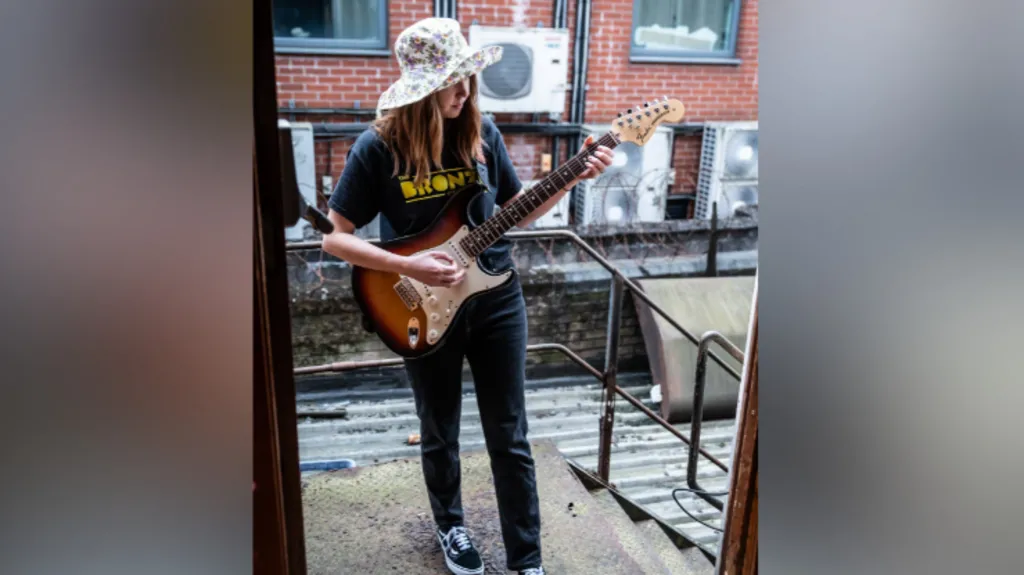 Sonja Sleator is playing an electric guitar. It's brown and white. She's wearing a hat with a flower pattern, a top that says the Bronze on it and jeans and trainers.  A brick building is behind her. She's standing outside of a doorway. 
