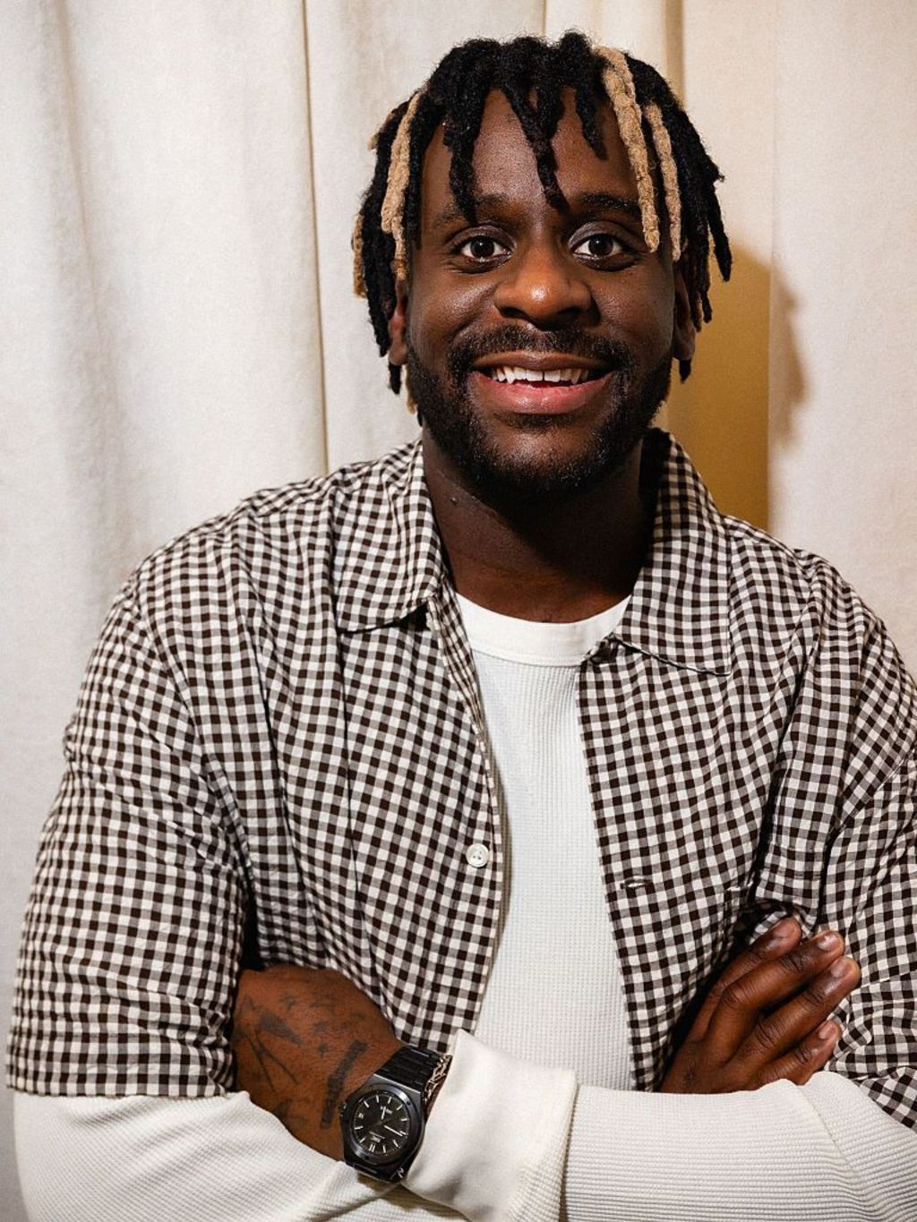 Myles Smith stands with his arms crossed in a white chequered shirt, while smiling.