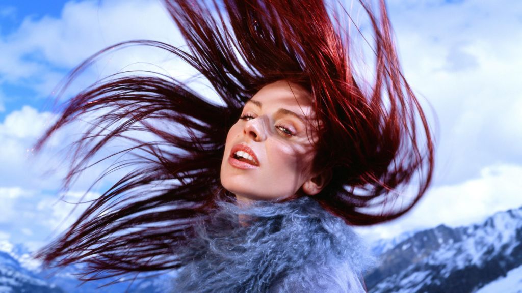 Rose Gray has red hair which she is sprawled in the air as she throws it back over her head. She is wearing a blue fluffy jacket. 